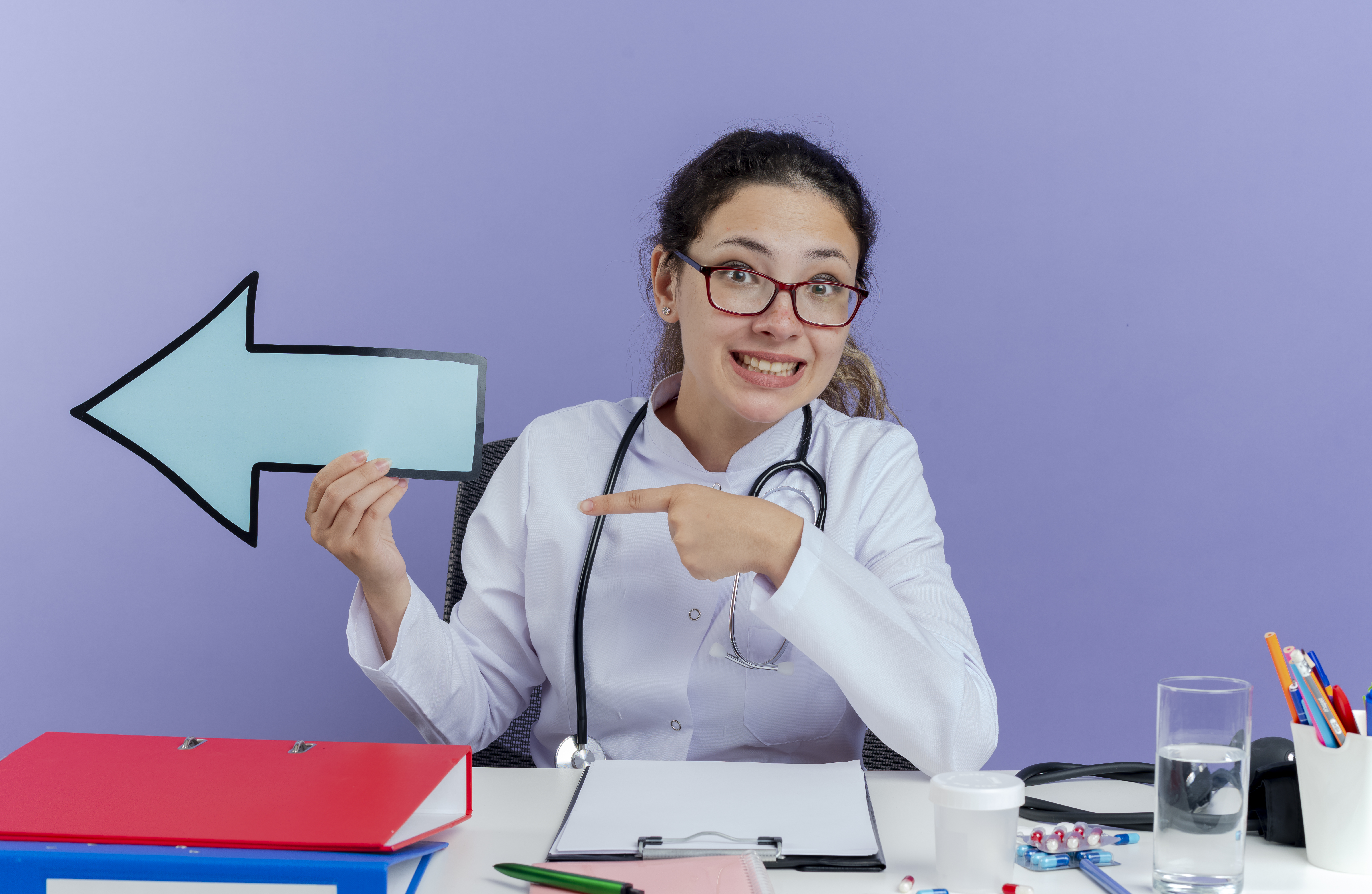 smiling young female doctor wearing medical robe and stethoscope sitting at desk with medical tools looking at camera holding arrow mark pointing at side isolated on purple background